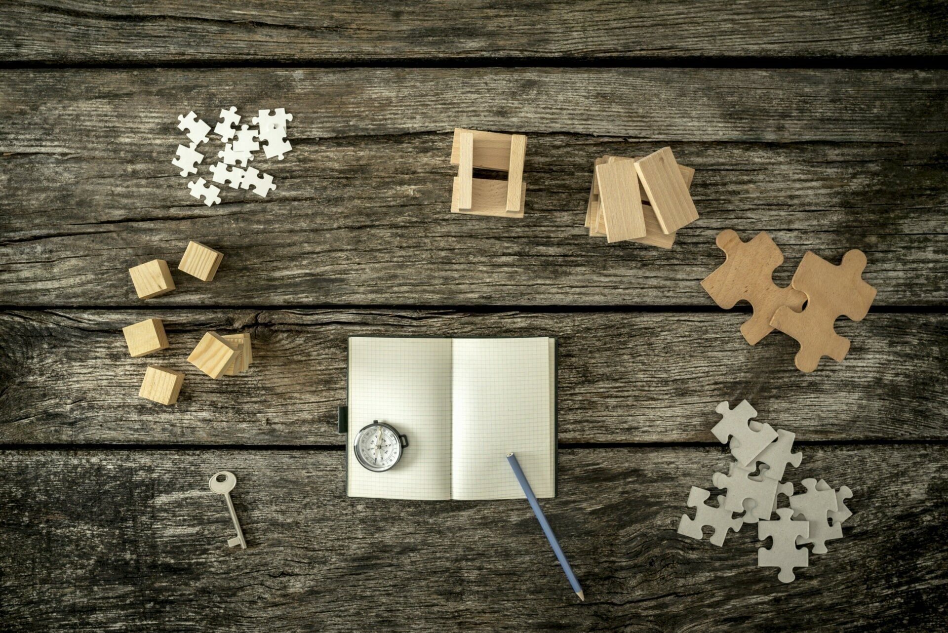 Various cubes, pegs, puzzles and a key lying on wooden desk arou Blick von oben auf vershciedene Puzzlteile, Klötze, einen Schlüssel, ein Notizblock, einen Kompass und einen Bleistift.