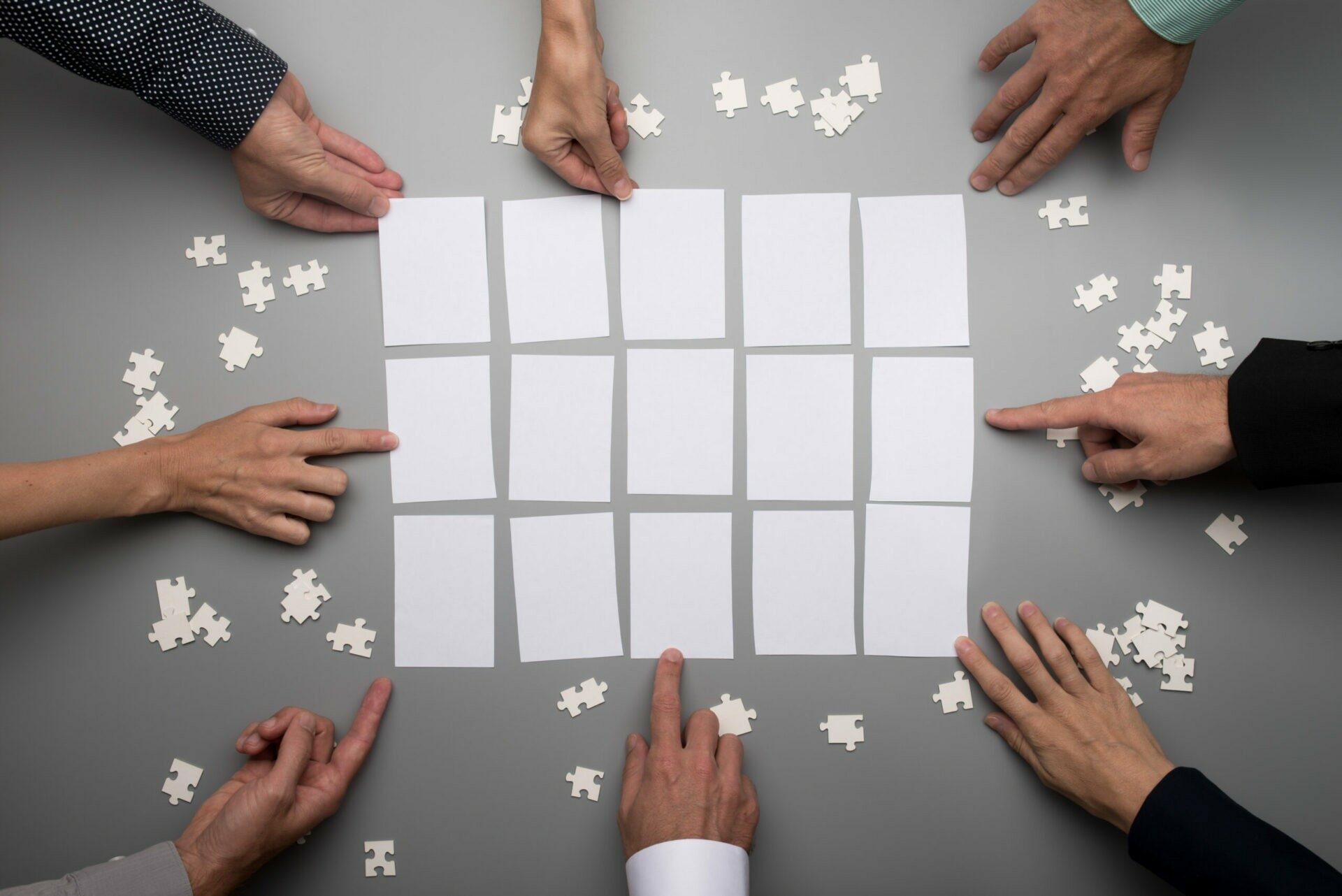 Top view of businesspeople hands touching empty white papers Fünfzehn Rechtecke die von Puzzleteilen umgeben sind. Acht verschiedene Hände greifen nach den Rechtecken.